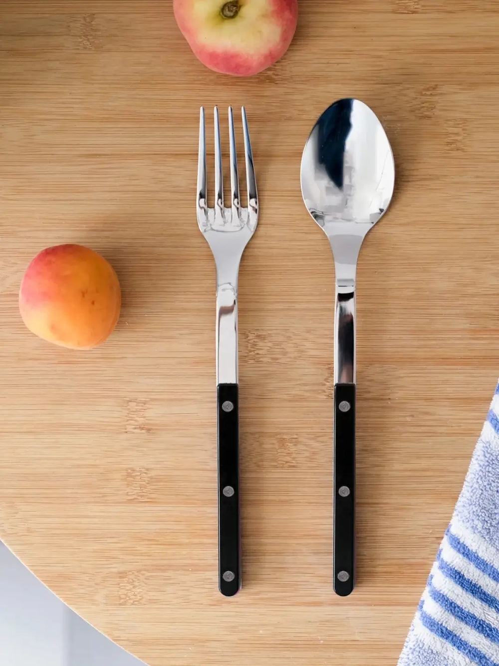 Shiny silver spoon and fork from Sabre Paris Bistrot Solid Shiny Serving Set in Black laid out on a light wood cutting board alongside fresh peaches and a blue-striped towel | Twentyseven Toronto