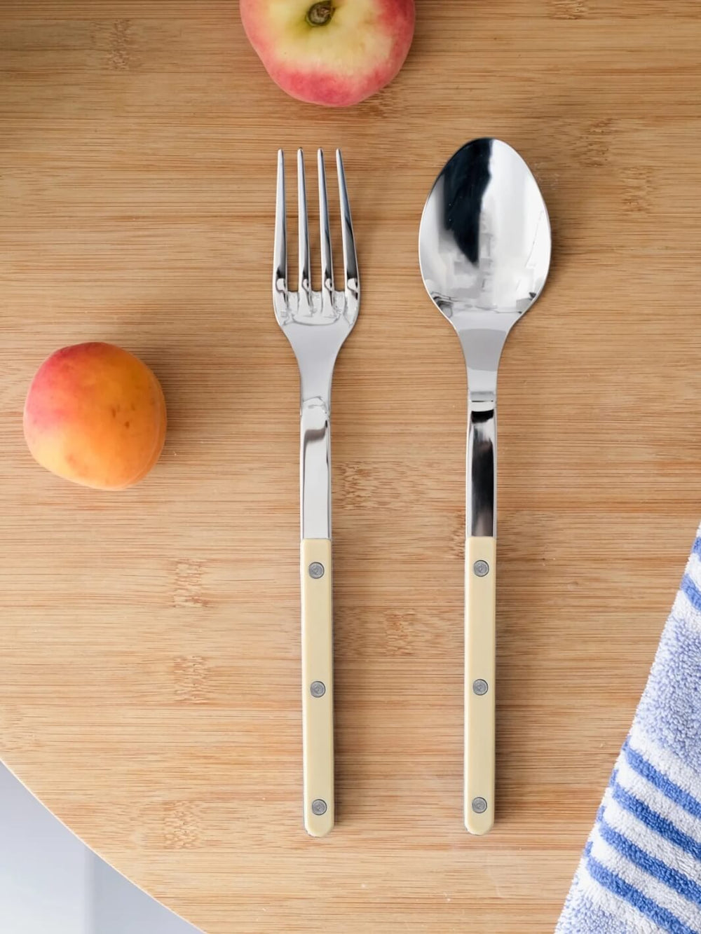 Shiny silver spoon and fork from Sabre Paris Bistrot Solid Shiny Serving Set in Ivory laid out on a light wood cutting board alongside fresh peaches and a blue-striped towel | Twentyseven Toronto