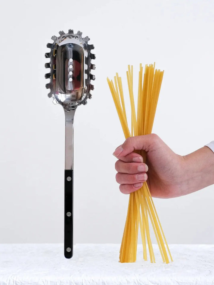 Sabre Paris Bistrot Solid Shiny Spaghetti Spoon in Black stands vertically on a textured white surface next to a hand holding a bundle of dry spaghetti | Twentyseven Toronto