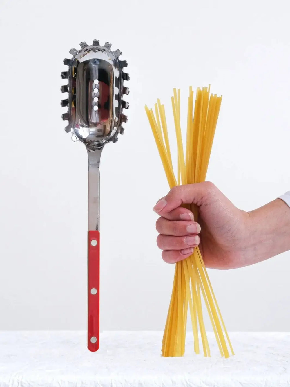 Sabre Paris Bistrot Solid Shiny Spaghetti Spoon in Burgundy stands vertically on a textured white surface next to a hand holding a bundle of dry spaghetti | Twentyseven Toronto