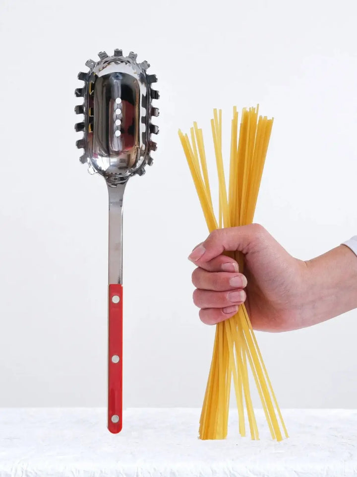 Sabre Paris Bistrot Solid Shiny Spaghetti Spoon in Burgundy stands vertically on a textured white surface next to a hand holding a bundle of dry spaghetti | Twentyseven Toronto