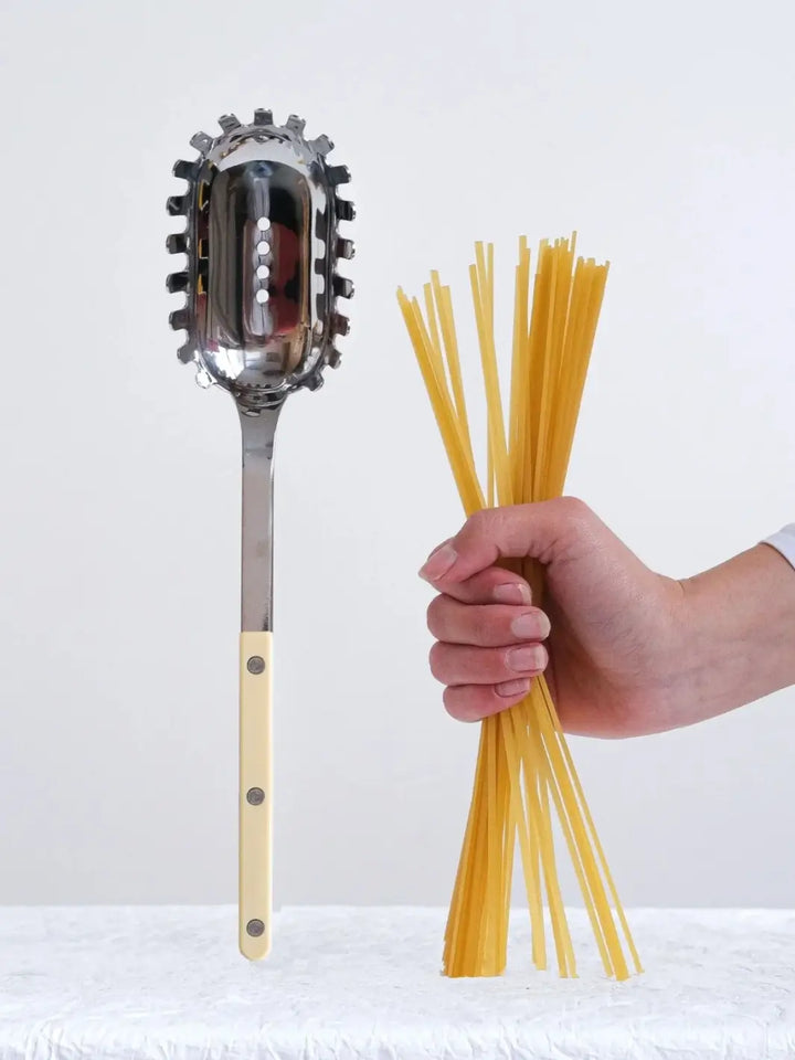 Sabre Paris Bistrot Solid Shiny Spaghetti Spoon in Ivory stands vertically on a textured white surface next to a hand holding a bundle of dry spaghetti | Twentyseven Toronto