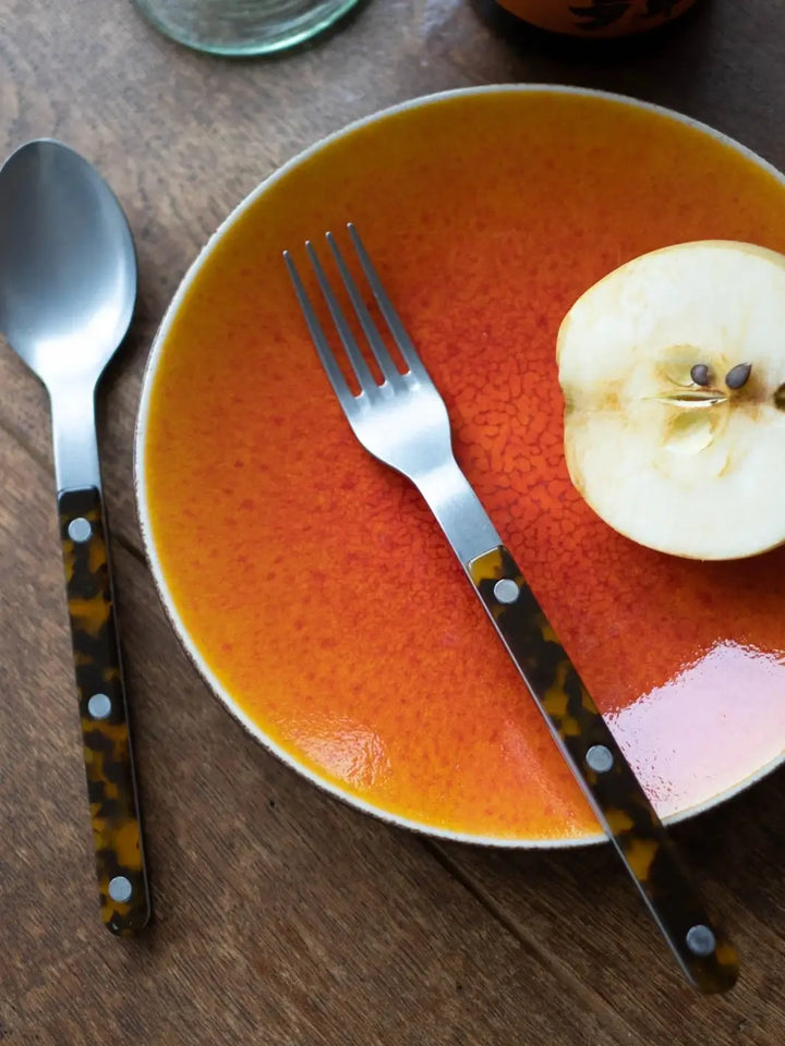 A dinner fork and soup spoon from the Sabre Paris Bistrot Vintage Finish 5 Piece Cutlery Set in Faux Tortoise arranged on a wooden surface next to an orange speckled plate containing half of an apple | Twentyseven Toronto