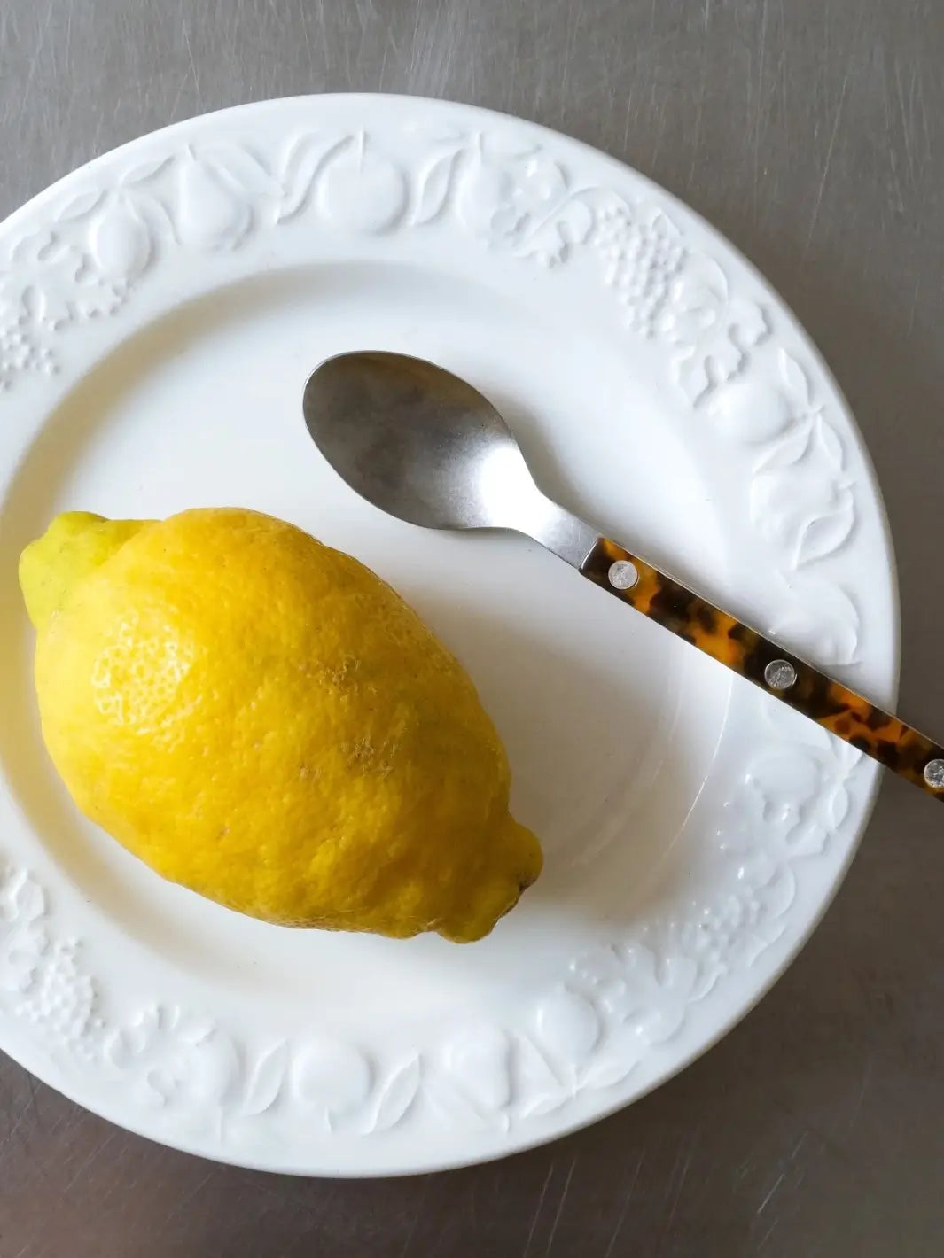 A teaspoon from the Sabre Paris Bistrot Vintage Finish 5 Piece Cutlery Set in Faux Tortoise resting on a white embossed ceramic plate alongside a yellow lemon | Twentyseven Toronto