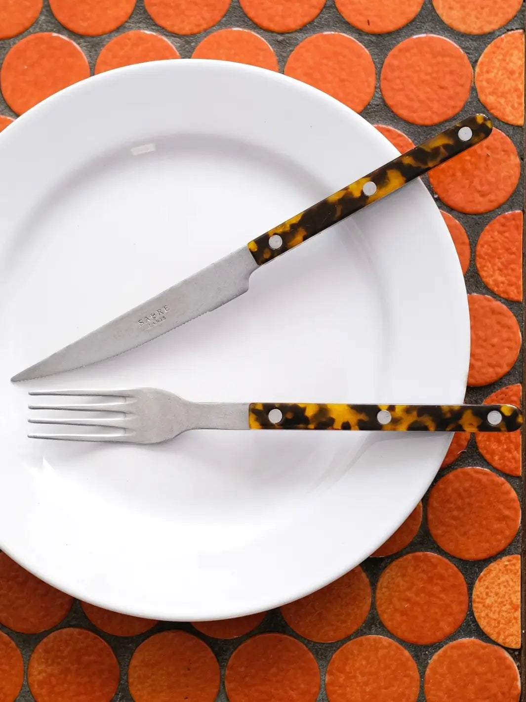 Dinner knife and soup spoon from the Sabre Paris Bistrot Vintage Finish 5 Piece Cutlery Set in Faux Tortoise are displayed on a white plate, against a background of bright orange circular tiles | Twentyseven Toronto