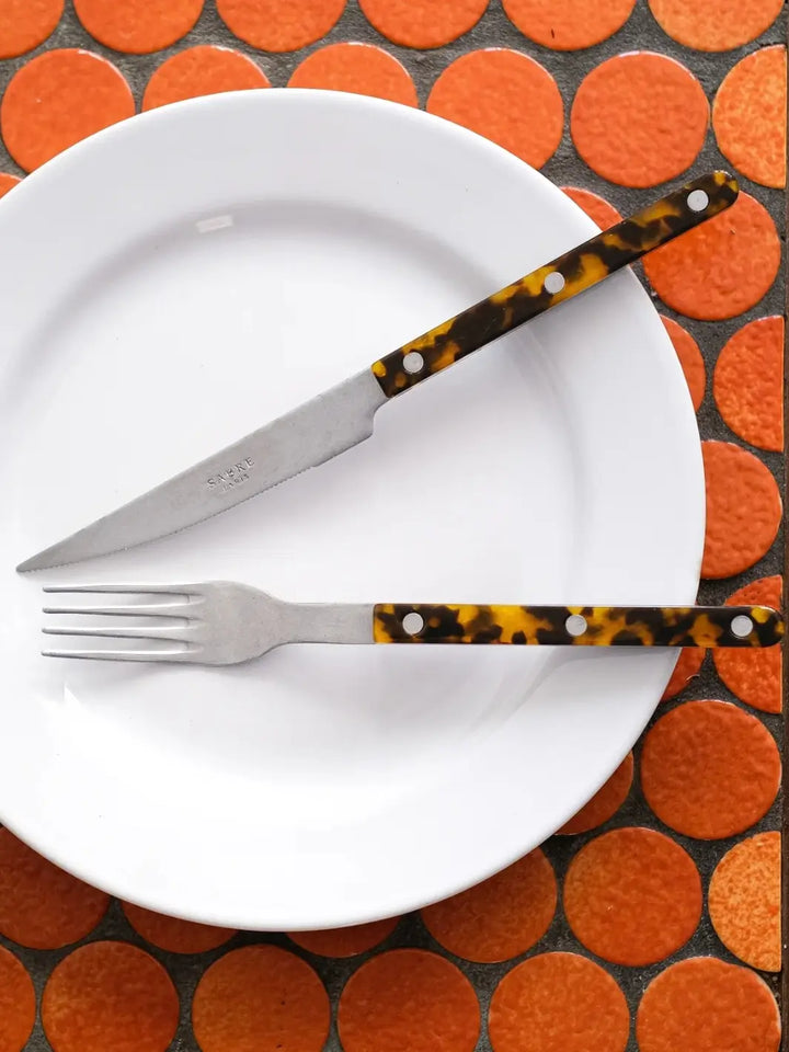 Dinner knife and soup spoon from the Sabre Paris Bistrot Vintage Finish 5 Piece Cutlery Set in Faux Tortoise are displayed on a white plate, against a background of bright orange circular tiles | Twentyseven Toronto