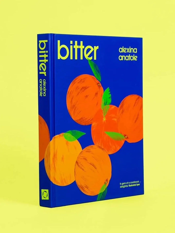 The hardcover cookbook "Bitter by Alexina Anatole" is displayed at an angle, showing a bold blue background illustrated with bright orange citrus fruits against a vivid yellow backdrop | Twentyseven Toronto