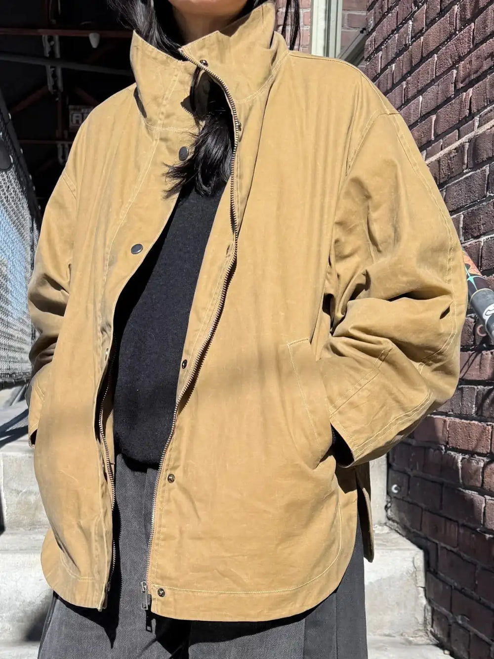 Person wearing a brown 'Blair Waxed Cotton Jacket' in 'Khaki' from Le Bon Shoppe standing against a brick wall | Twentyseven Toronto