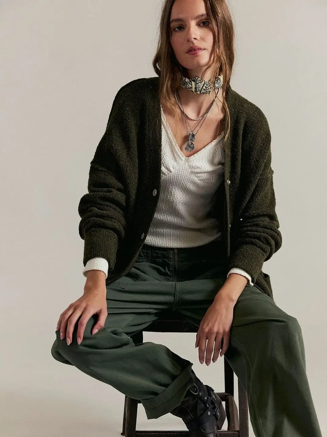 A model sits on a stool wearing the open Free People Blazing Cardi in Dark Green styled over a white top and olive green trousers | Twentyseven Toronto
