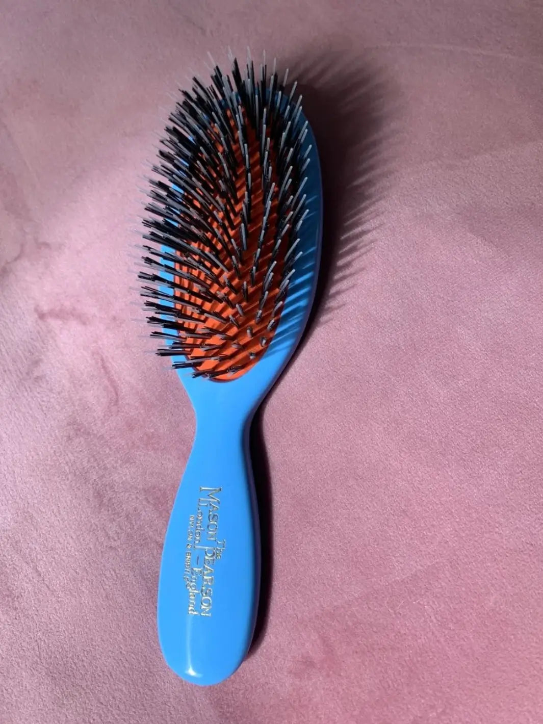 A premium baby blue hairbrush with mixed black bristles on a bright orange rubber cushion is showcased against a soft pink velvet backdrop, highlighting the luxury design of the Mason Pearson Baby Blue Boar Bristle & Nylon Mix Hair Brush | Twentyseven Toronto