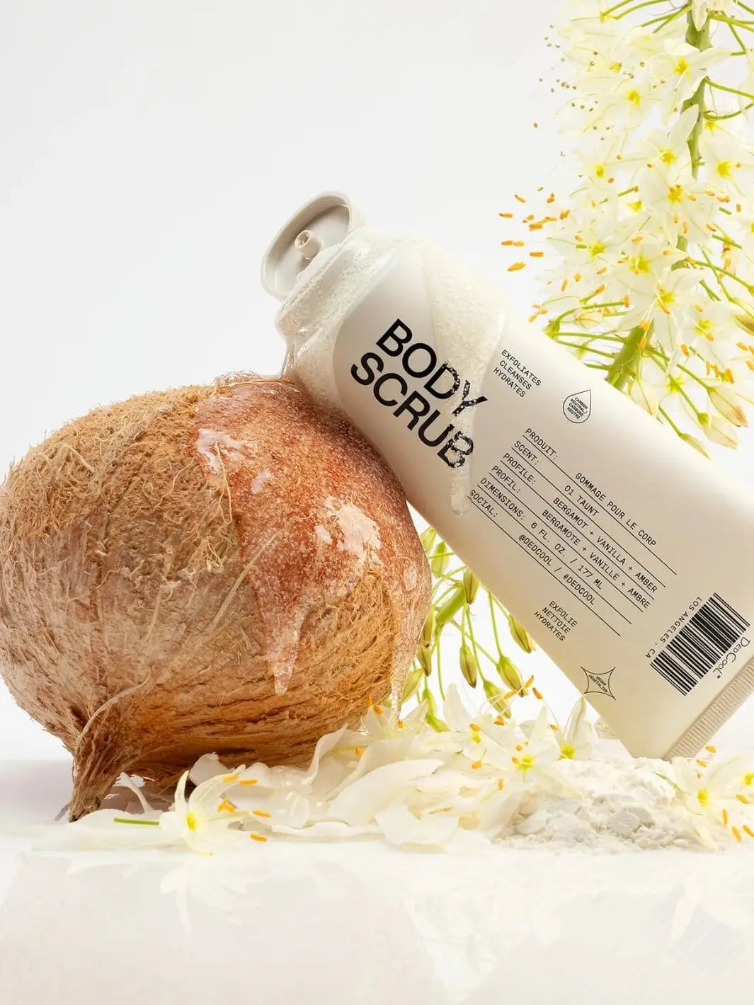 A squeeze tube of Dedcool Body Scrub Xtra Milk rests a coconut on a bed of white flowers | Twentyseven Toronto