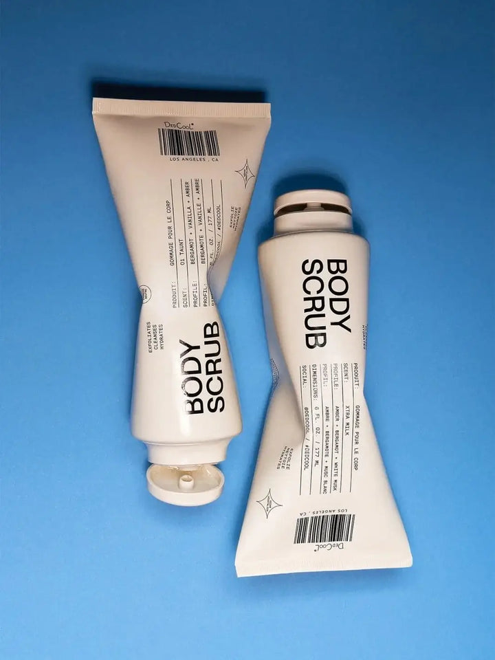 Two white squeeze tubes of Dedcool Body Scrub Xtra Milk are positioned side by side facing opposite directions on a bright blue background | Twentyseven Toronto