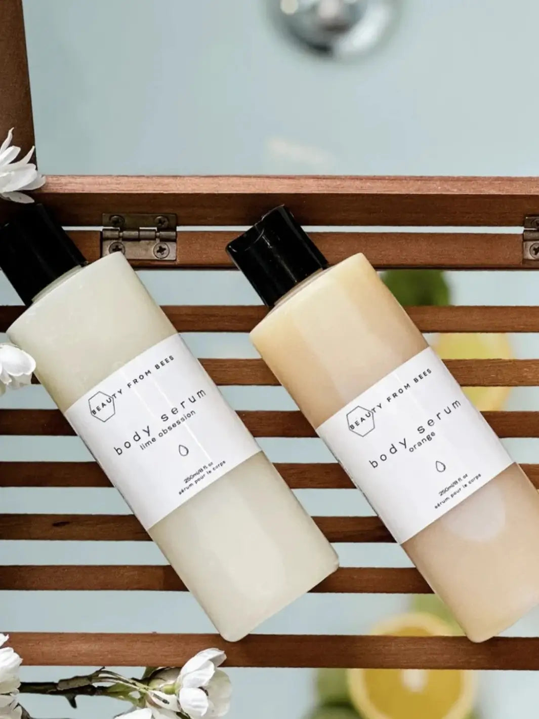 Two bottles of Beauty from Bees Body Serum in Lime Obsession and Orange resting side-by-side on a wooden bath tray | Twentyseven