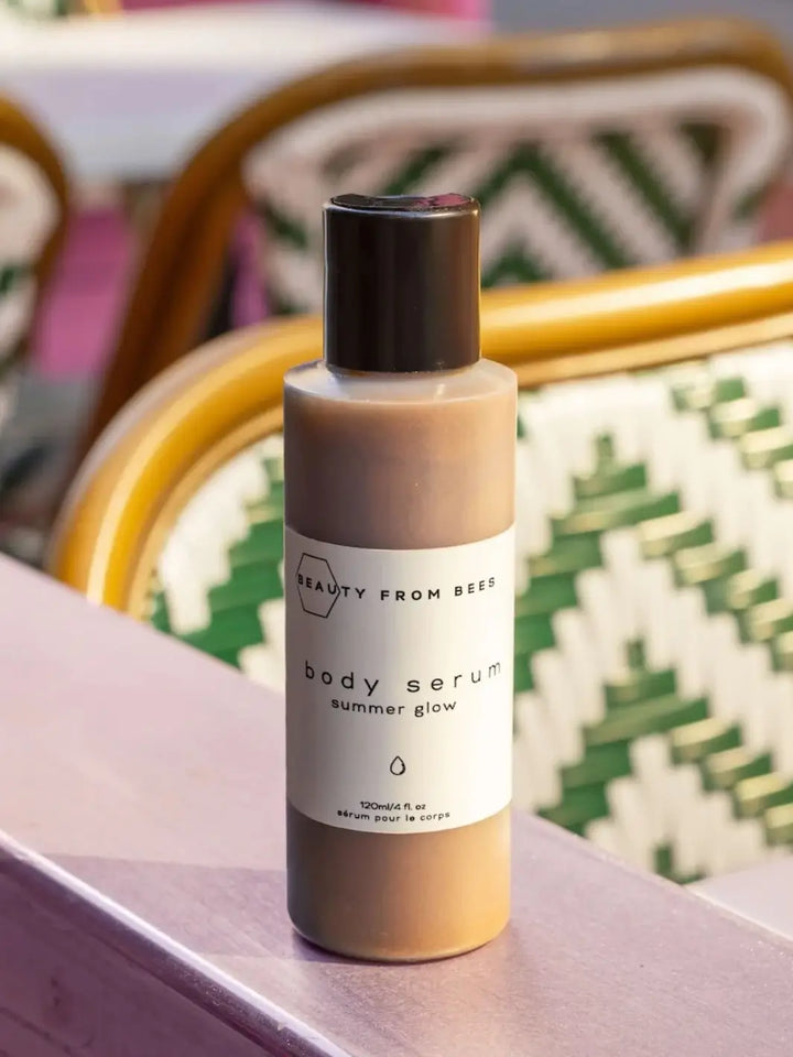 A bottle of Beauty from Bees Body Serum - Summer Glow stands on a wooden ledge with a blurred background of green and white patterned outdoor bistro chairs | Twentyseven Toronto