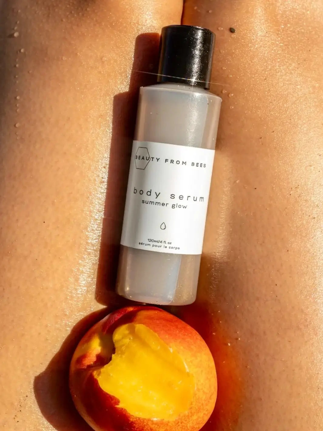 A bottle of Beauty from Bees Body Serum - Summer Glow rests between a person's sunlit legs next to a ripe, cut peach on a beach | Twentyseven Toronto