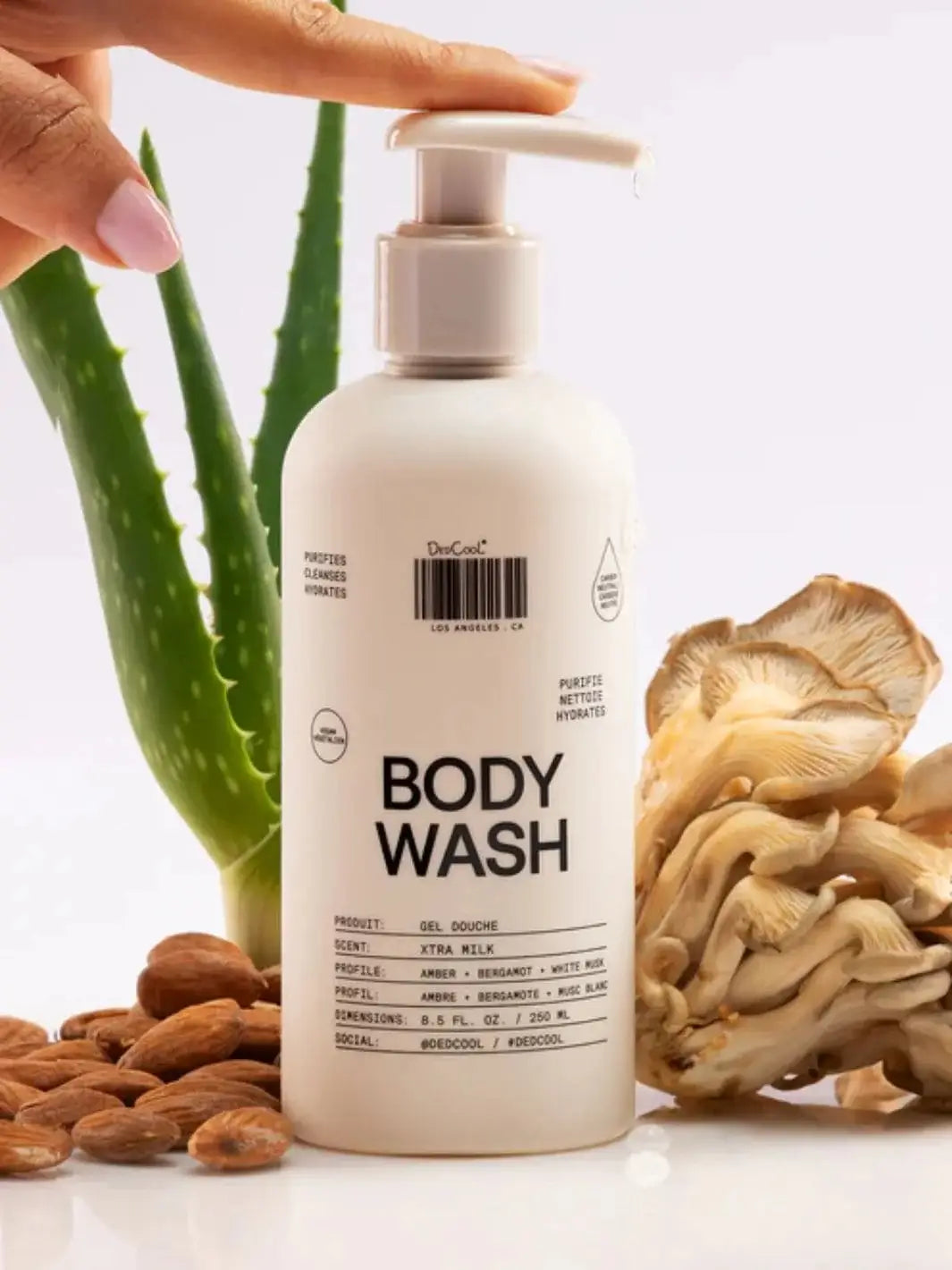 A hand presses the pump of a white Dedcool Body Wash Xtra Milk bottle arranged on a bright surface with almonds, an aloe vera plant, and oyster mushrooms | Twentyseven Toronto