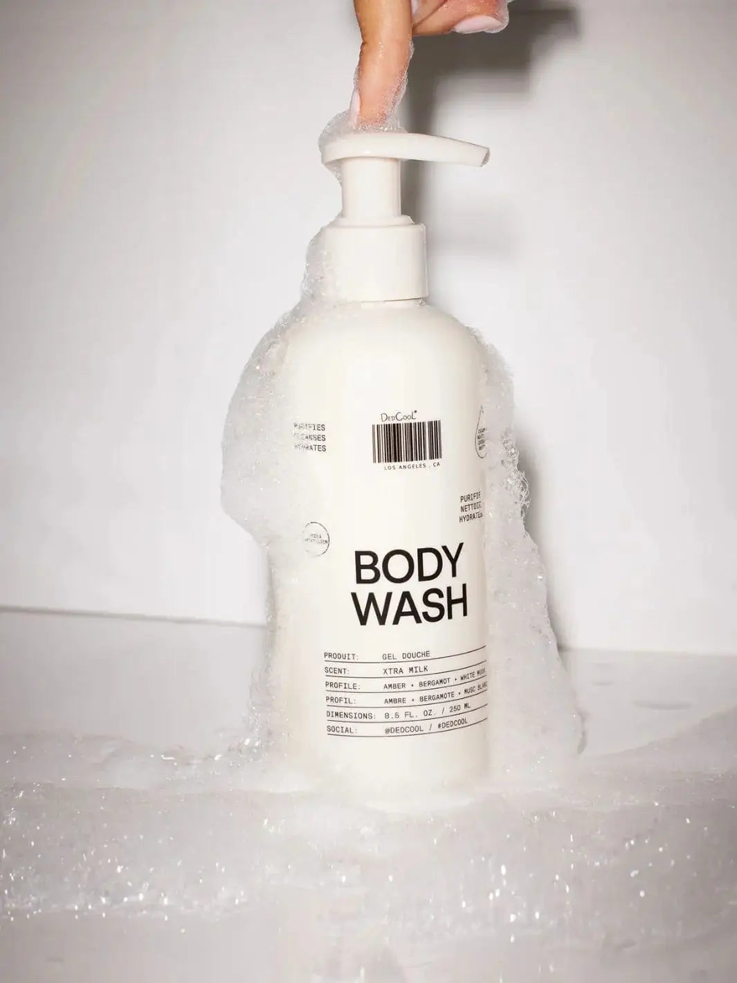 A finger presses the top of a white Dedcool Body Wash Xtra Milk bottle that is covered in a thick layer of white soap suds and bubbles | Twentyseven Toronto