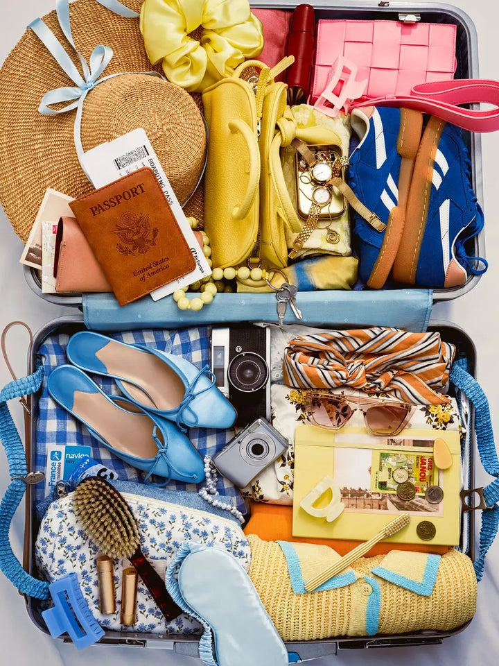 A perfectly packed suitcase filled with colorful travel essentials like a passport, blue shoes, sunglasses, and cameras forms the vibrant artwork for the Piecework Puzzles Bon Voyage 1000 Piece Puzzle | Twentyseven Toronto