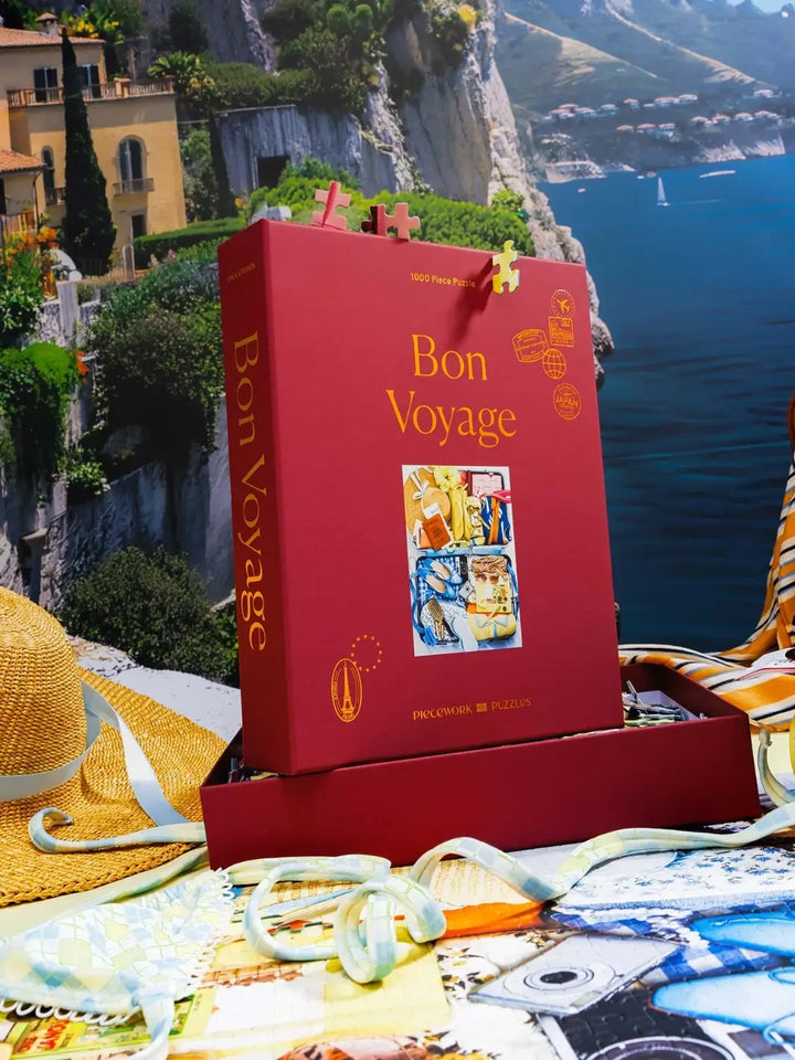 The deep red box of the Piecework Puzzles Bon Voyage 1000 Piece Puzzle stands upright on top of a partially assembled jigsaw puzzle against a scenic coastal backdrop | Twentyseven Toronto