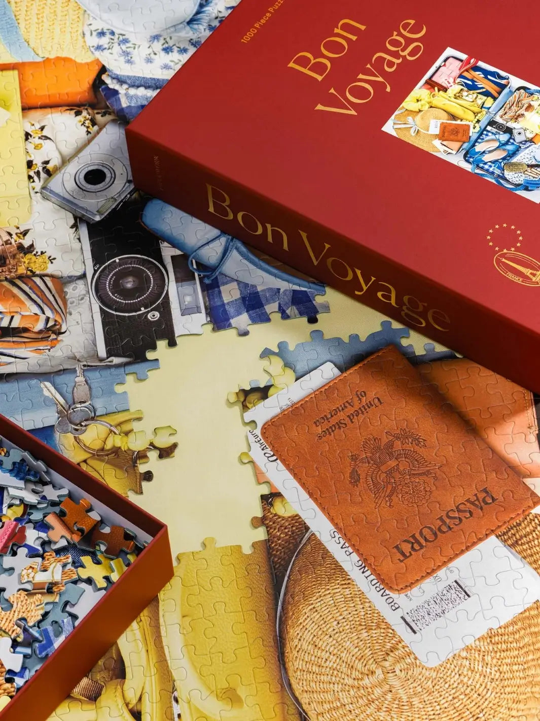 The red cover of the Piecework Puzzles Bon Voyage 1000 Piece Puzzle lies across an in-progress jigsaw puzzle next to a brown passport holder containing a boarding pass | Twentyseven Toronto