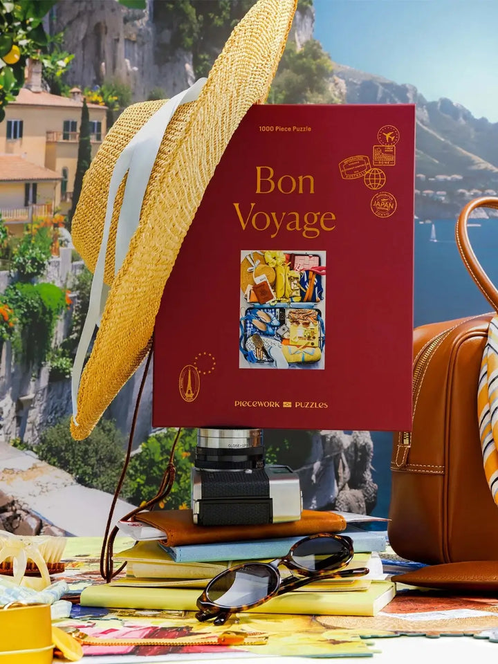 The Piecework Puzzles Bon Voyage 1000 Piece Puzzle box is styled vertically on top of stacked books and a camera, adorned with a straw sun hat and tortoise shell sunglasses | Twentyseven Toronto