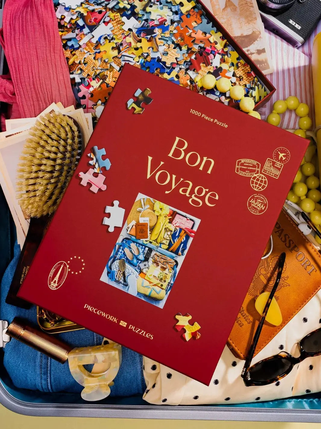 The red box of the Piecework Puzzles Bon Voyage 1000 Piece Puzzle is styled flat inside an open suitcase surrounded by travel accessories, a hairbrush, and loose puzzle pieces | Twentyseven Toronto
