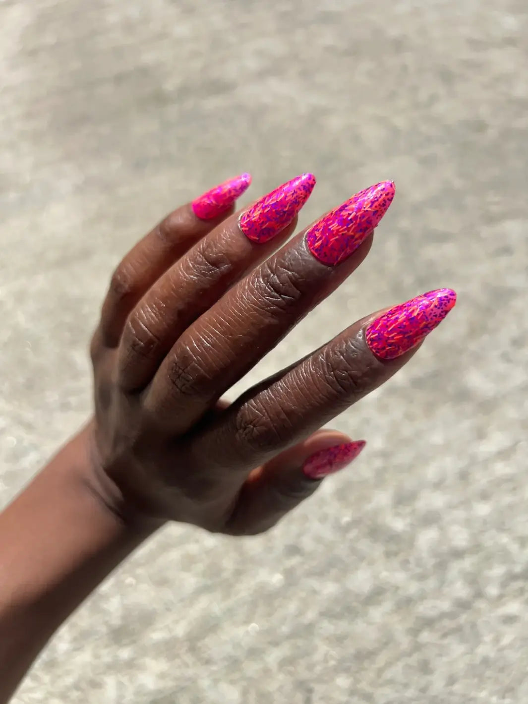 A close-up shows a hand with a deep skin tone and long, almond-shaped fingernails painted with bright pink Death Valley Nails Bone Daddy Nail Polish against a blurry outdoor background | Twentyseven Toronto