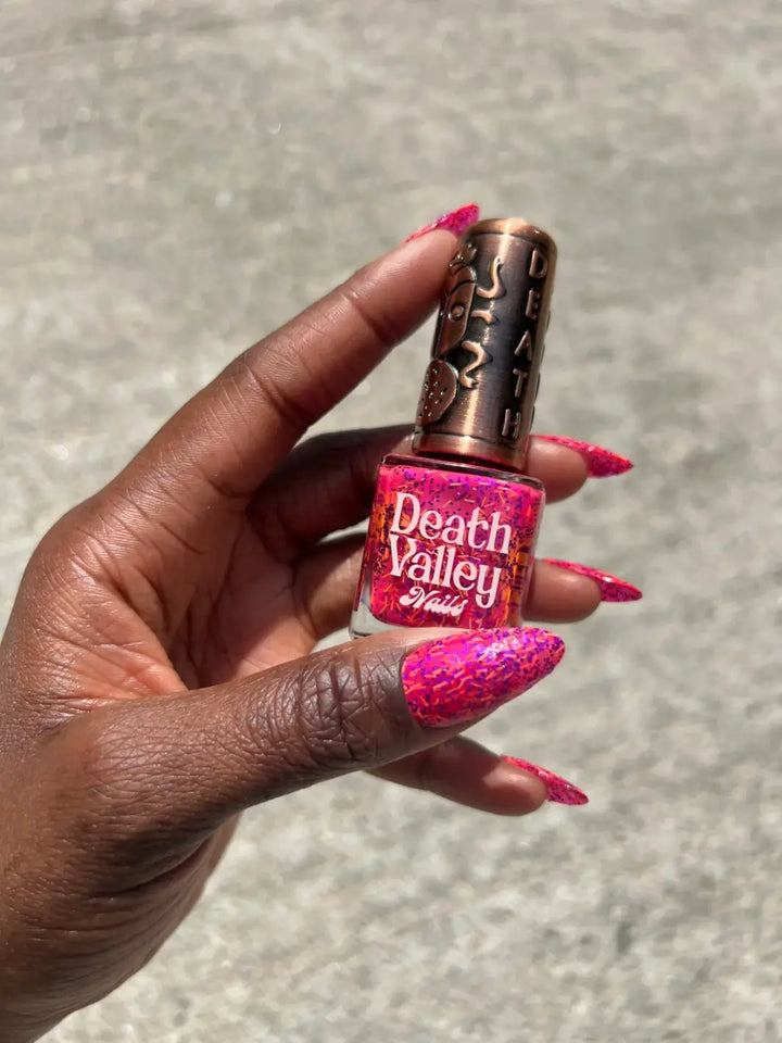 A hand with a deep skin tone holds a glass bottle of Death Valley Nails Bone Daddy Nail Polish, matching the neon pink lacquer inside with a manicure on long, stiletto nails | Twentyseven Toronto