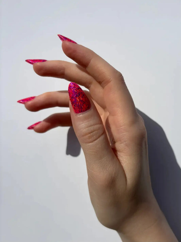A hand with a fair skin tone and long, stiletto nails is against a plain white background, showcasing the vibrant bright pink shade of Death Valley Nails Bone Daddy Nail Polish | Twentyseven Toronto