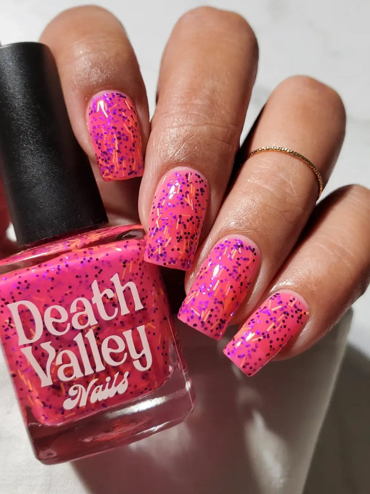 A hand with long, square-shaped fingernails wearing bright pink Death Valley Nails Bone Daddy Nail Polish holding its glass bottle | Twentyseven Toronto
