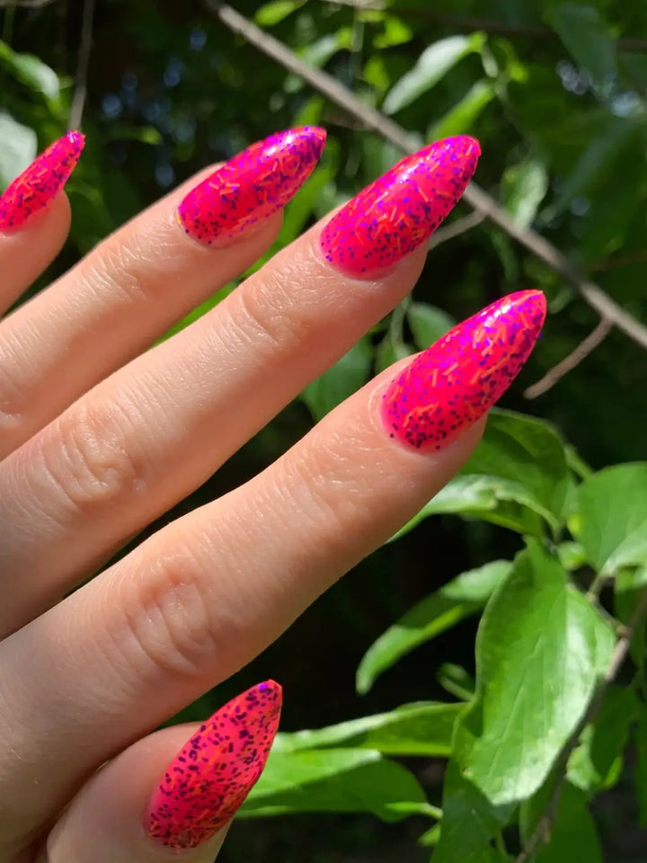 A hand with long, almond-shaped bright pink nails is spread out against a lush green leafy background to show off a full manicure of Death Valley Nails Bone Daddy Nail Polish | Twentyseven Toronto
