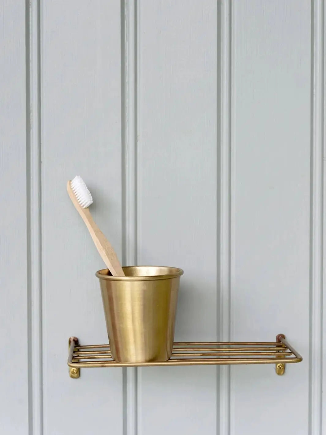 A wooden toothbrush rests inside a Fog Linen Work Brass Cup sitting on a matching wire shelf against a grey wood paneled wall | Twentyseven Toronto