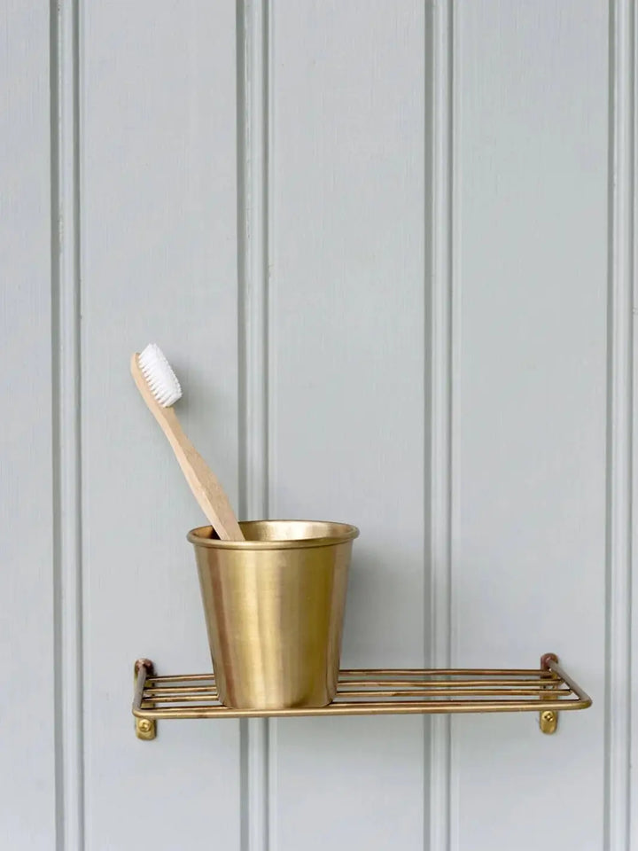 A wooden toothbrush rests inside a Fog Linen Work Brass Cup sitting on a matching wire shelf against a grey wood paneled wall | Twentyseven Toronto