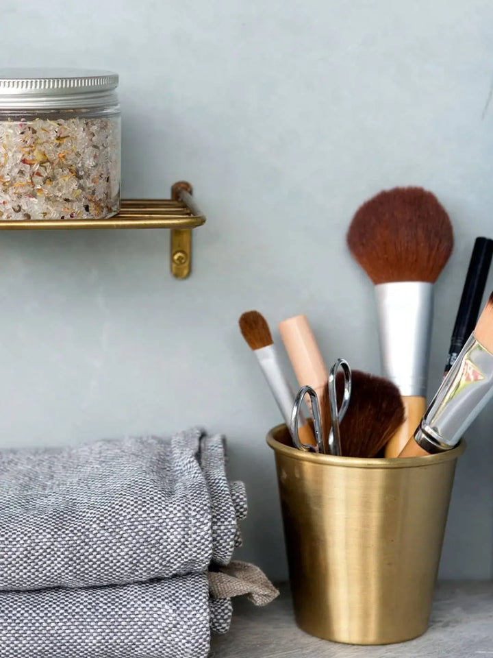 A Fog Linen Work Brass Cup filled with makeup brushes and grooming tools stands next to folded grey towels in a bathroom setting | Twentyseven Toronto