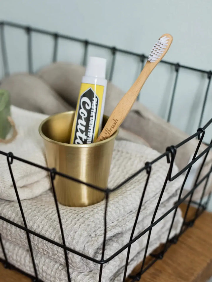 A wooden toothbrush and toothpaste tube are stored inside a Fog Linen Work Brass Cup nestled within a black wire basket filled with neutral towels | Twentyseven Toronto