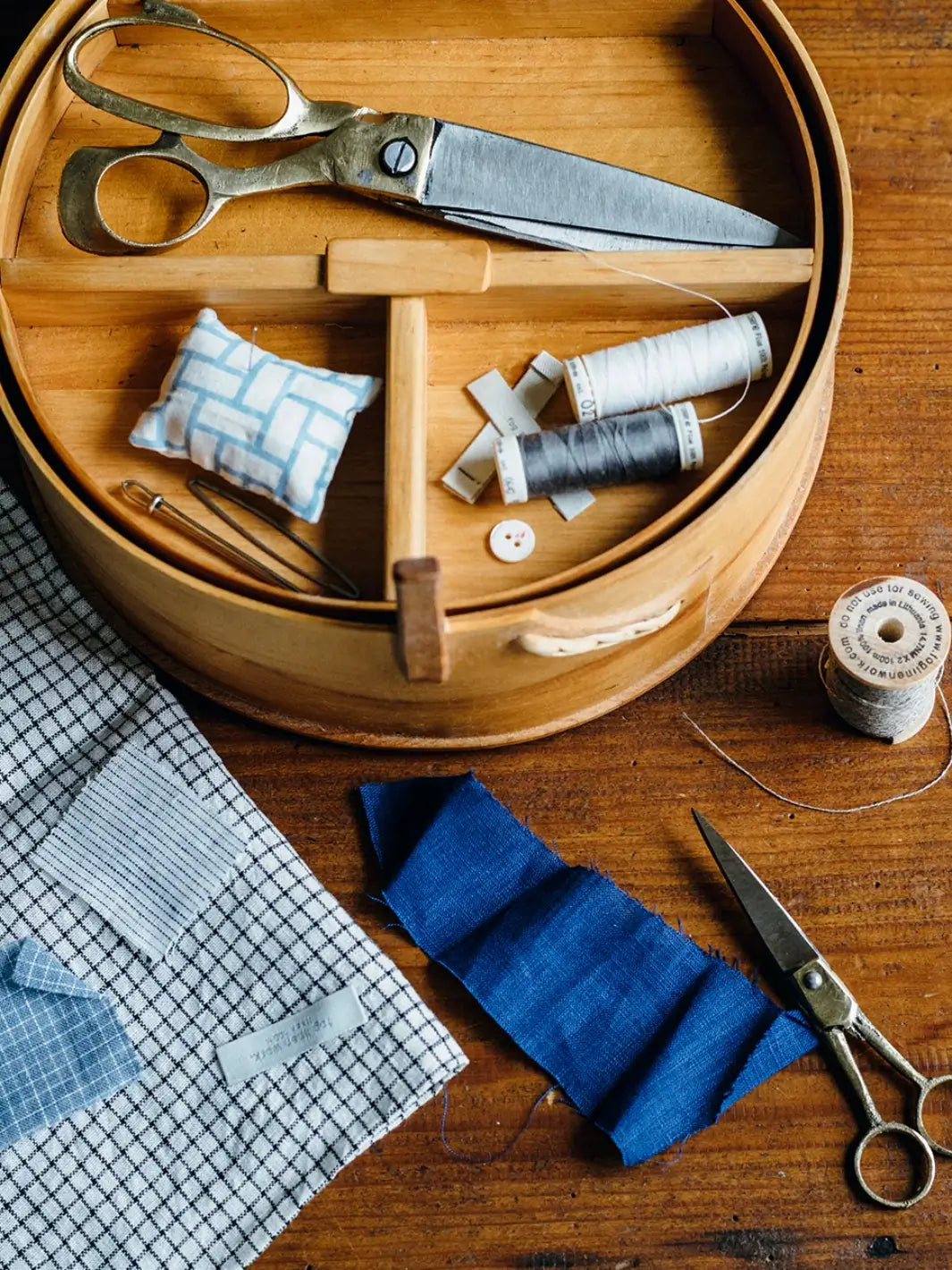 Brass Handle Scissors Small from Fog Linen Work sit on a wooden tabletop next to a sewing basket and blue linen fabric scraps | Twentyseven Toronto