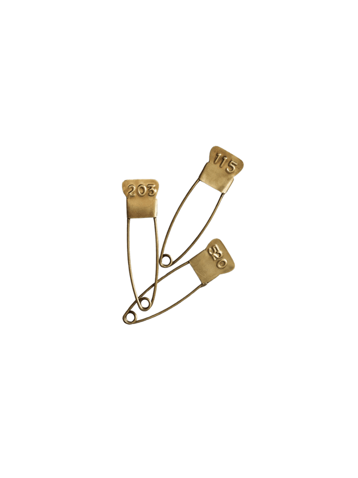 A set of three vintage-inspired Brass Number Pin from Fog Linen Work fasteners, displaying different three-digit codes on their antique-style heads | Twentyseven Toronto