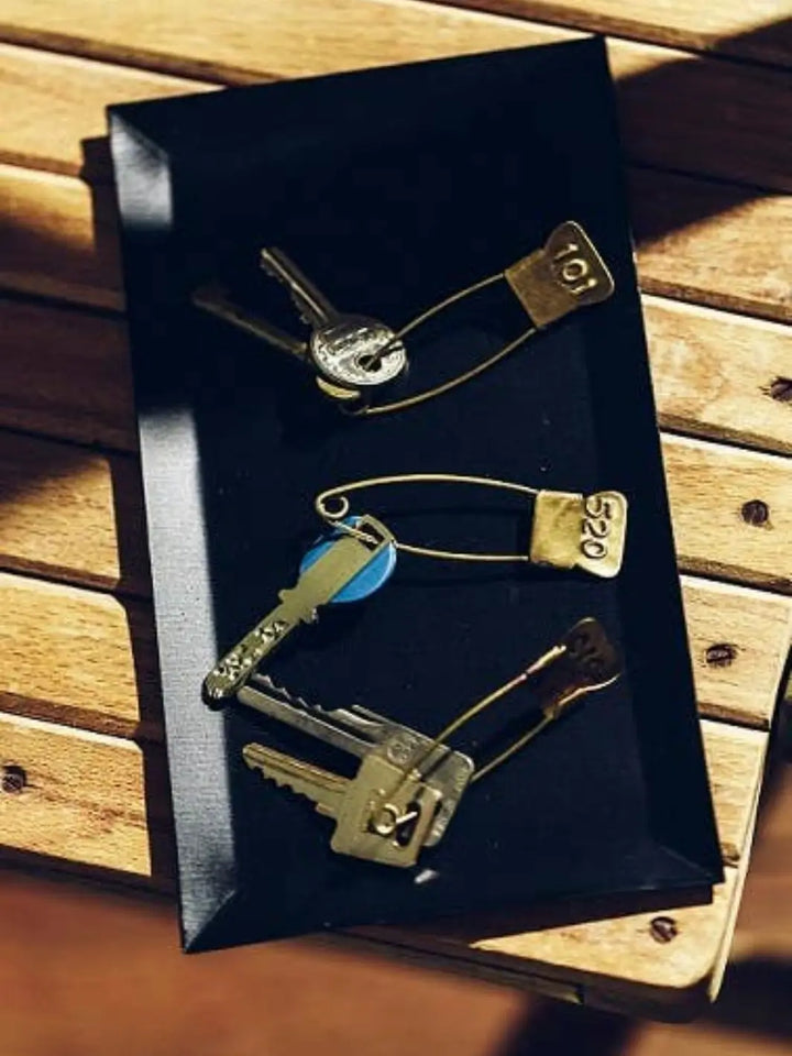 A sleek black tray holds several keys organized by the Brass Number Pin from Fog Linen Work, featuring stamped numerical identification tags | Twentyseven Toronto