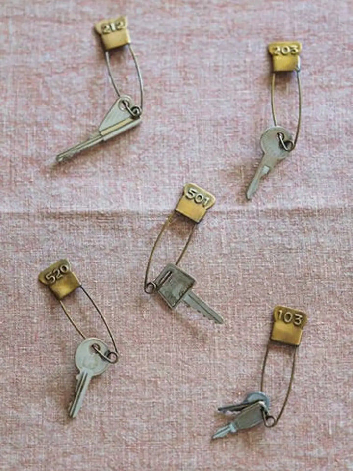 Five individual keys are spread across a textured pink fabric, each secured by a Brass Number Pin from Fog Linen Work with unique engraved numbers | Twentyseven Toronto