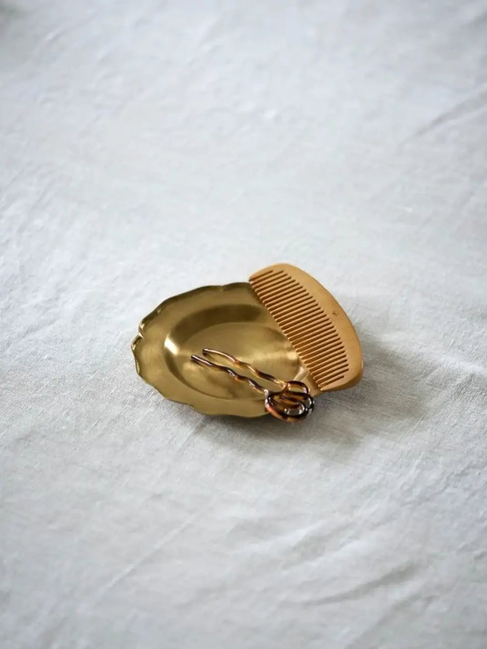 Fog Linen Work Brass Ruffle Edge Tray Small is displayed on a light grey textured fabric, holding a small tortoiseshell hair pin alongside a wooden comb | Twentyseven Toronto