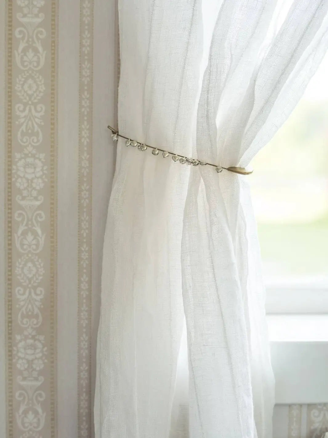 A sheer white linen curtain is elegantly held back by a Fog Linen Work Brass Safety Pin Bell Medium next to a patterned wall | Twentyseven Toronto