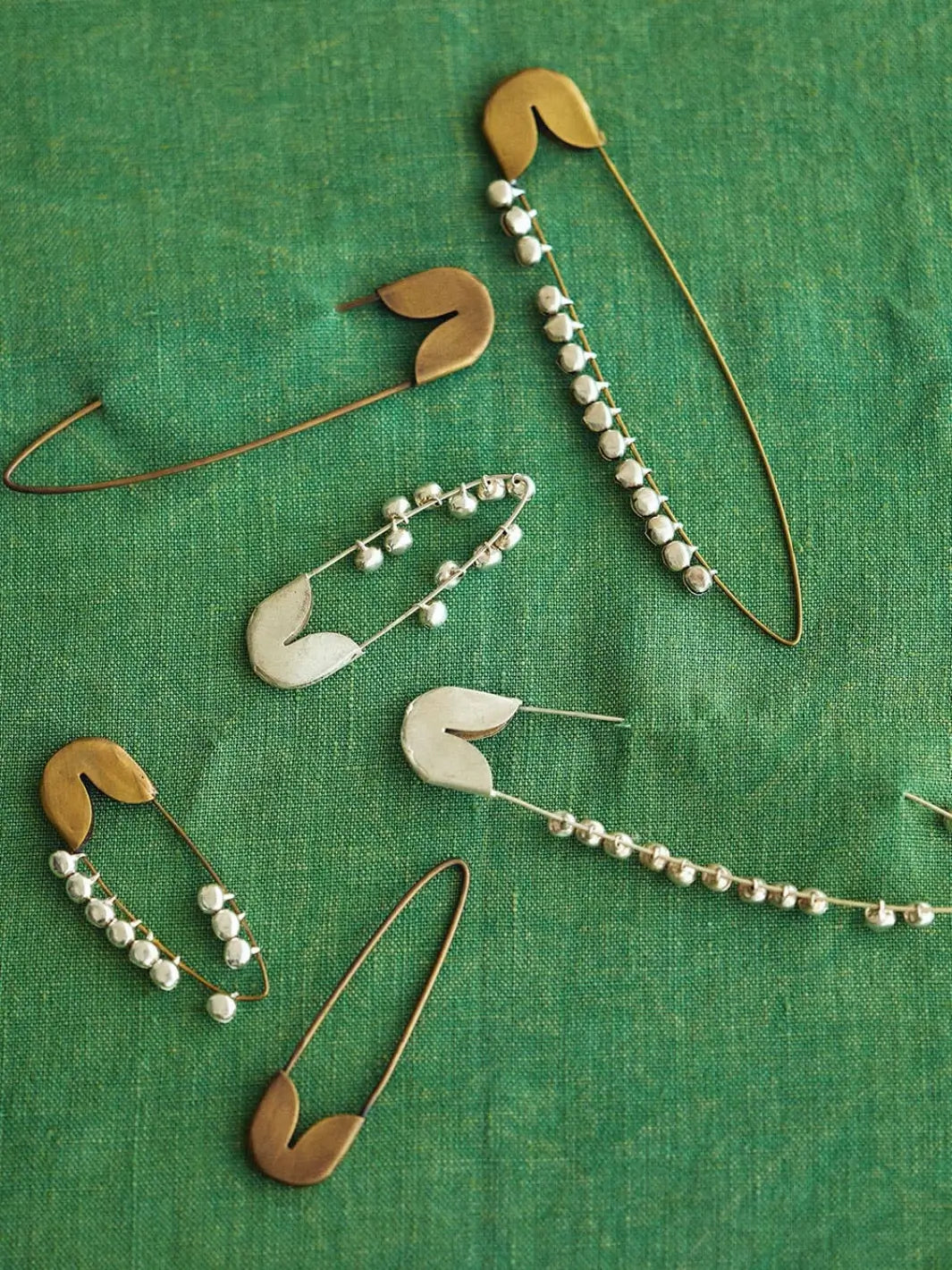 A collection of unique metal fasteners features a Fog Linen Work Brass Safety Pin Bell Medium displayed alongside other bell-adorned pins on a green textured fabric | Twentyseven Toronto