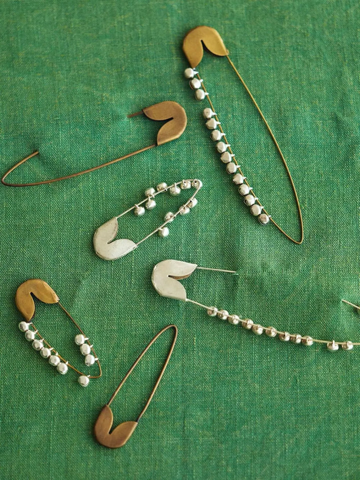 A collection of unique metal fasteners features a Fog Linen Work Brass Safety Pin Bell Medium displayed alongside other bell-adorned pins on a green textured fabric | Twentyseven Toronto