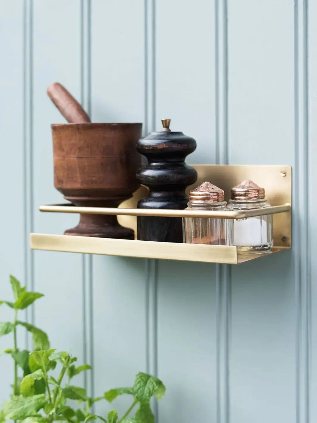 A Brass Shelf from Fog Linen Work is mounted on a light blue paneled wall, stylishly holding a wooden mortar and pestle along with vintage spice jars | Twentyseven Toronto