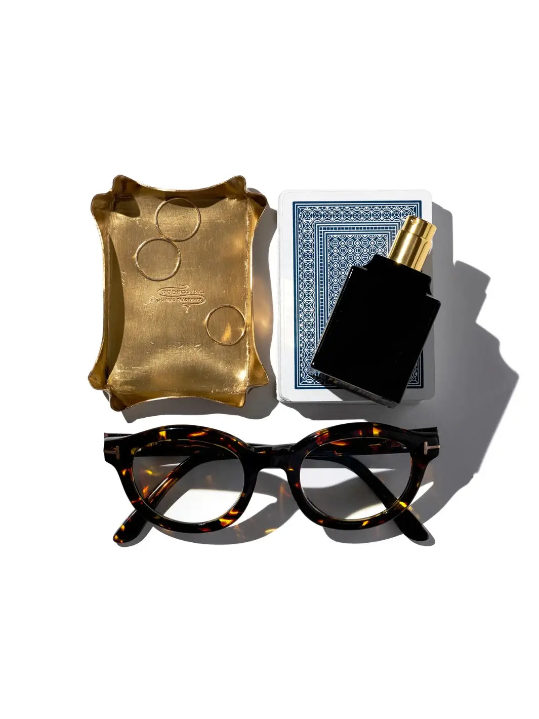 A gold Brass Small Tray from Puebco is neatly arranged alongside tortoiseshell glasses, a deck of playing cards, and a black perfume bottle on a white surface | Twentyseven Toronto