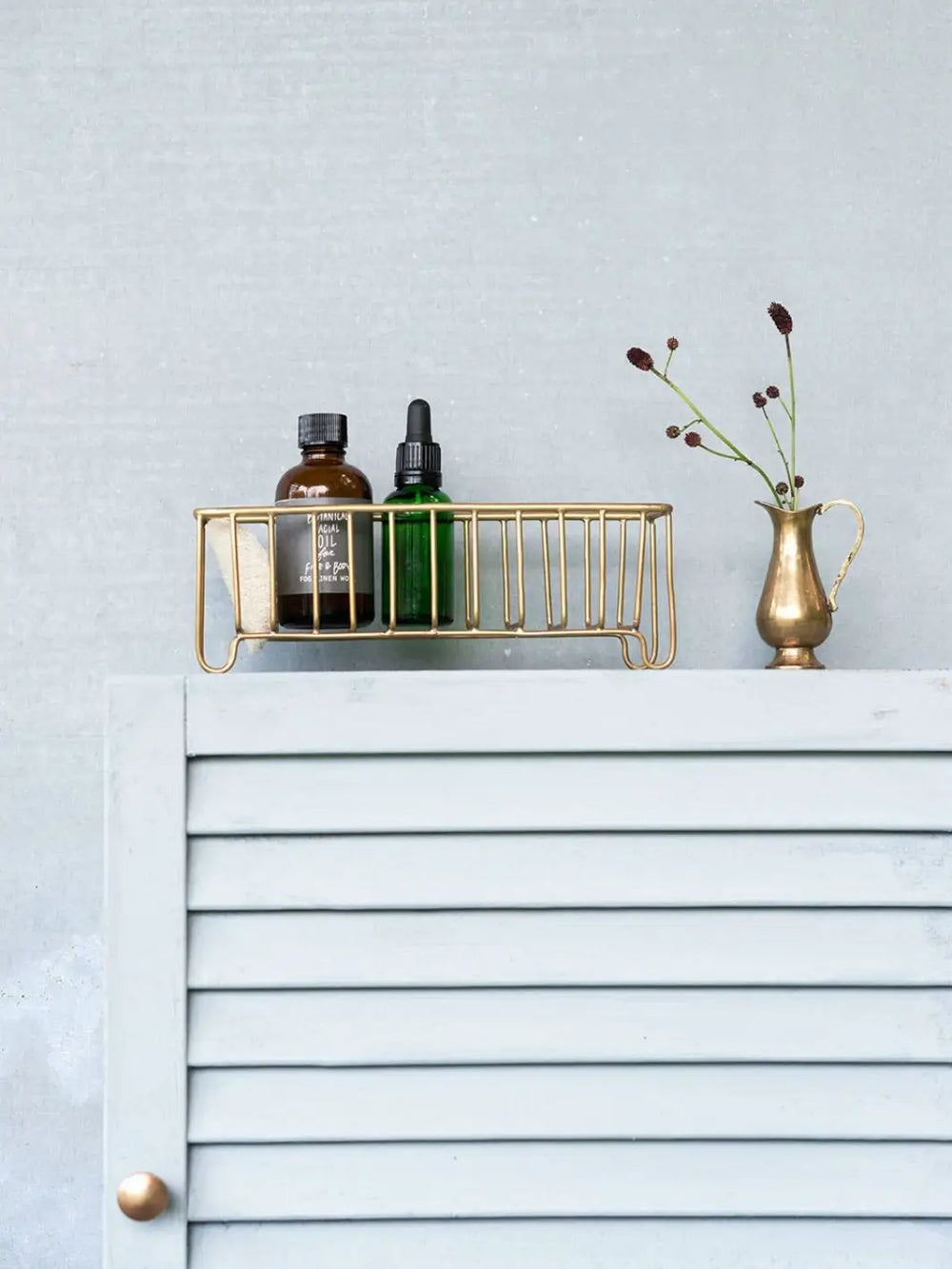 A Fog Linen Work Brass Wire Basket Narrow holds two small apothecary bottles on top of a light grey louvered cabinet next to a small brass vase | Twentyseven Toronto