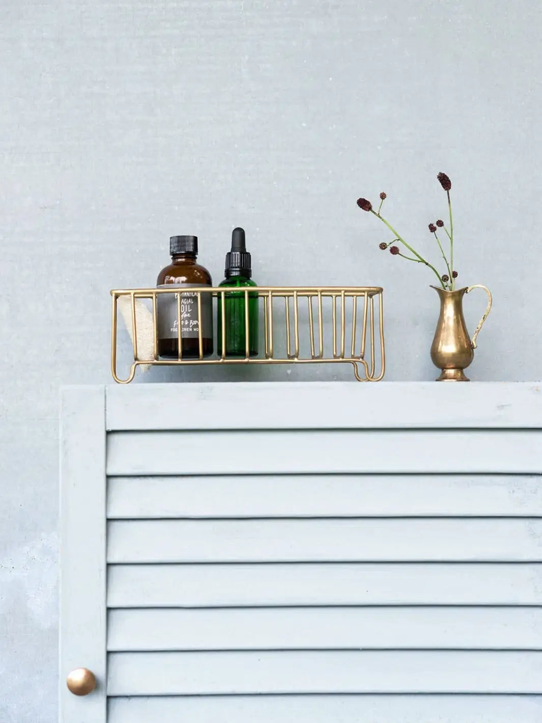 A Fog Linen Work Brass Wire Basket Narrow holds two small apothecary bottles on top of a light grey louvered cabinet next to a small brass vase | Twentyseven Toronto