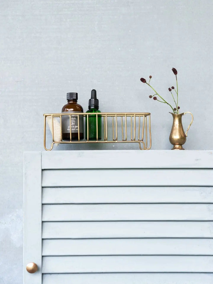 A Fog Linen Work Brass Wire Basket Narrow holds two small apothecary bottles on top of a light grey louvered cabinet next to a small brass vase | Twentyseven Toronto