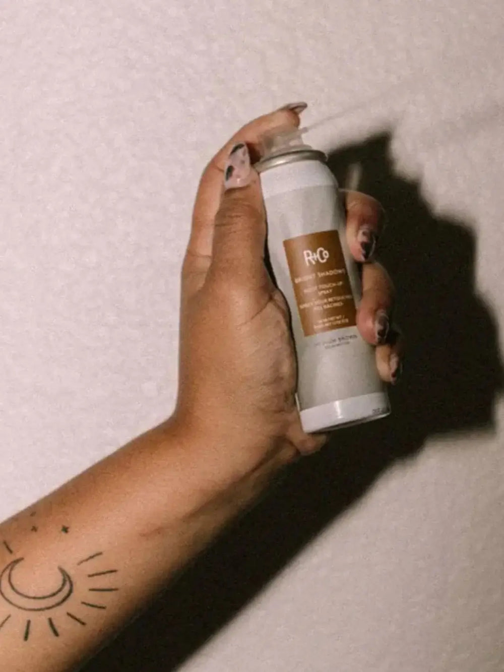 A hand with a crescent moon tattoo holds up an aerosol bottle of R+Co BRIGHT SHADOWS Root Touch Up Spray - Dark Brown against a neutral wall | Twentyseven Toronto