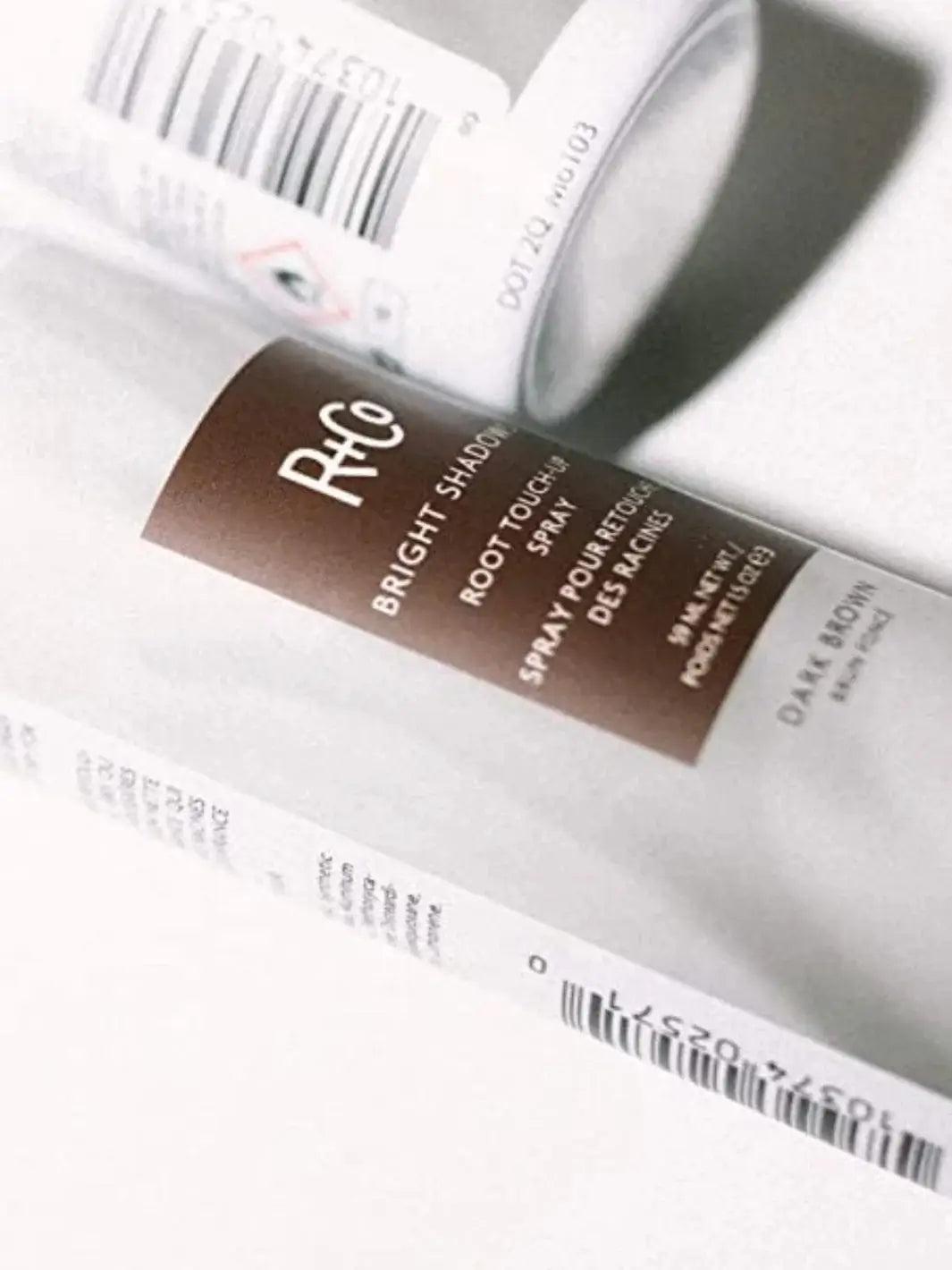 A close-up shot focuses on the sleek label details of an R+Co BRIGHT SHADOWS Root Touch Up Spray - Dark Brown bottle laying on a clean white surface | Twentyseven Toronto